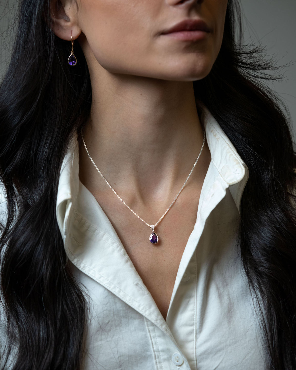 A close-up of a woman wearing a delicate white gold chain necklace with a teardrop-shaped purple gemstone pendant, and pear shaped amethyst dangles in yellow gold paired with a white collared shirt.