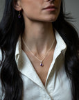 A close-up of a woman wearing a delicate white gold chain necklace with a teardrop-shaped purple gemstone pendant, and pear shaped amethyst dangles in yellow gold paired with a white collared shirt.