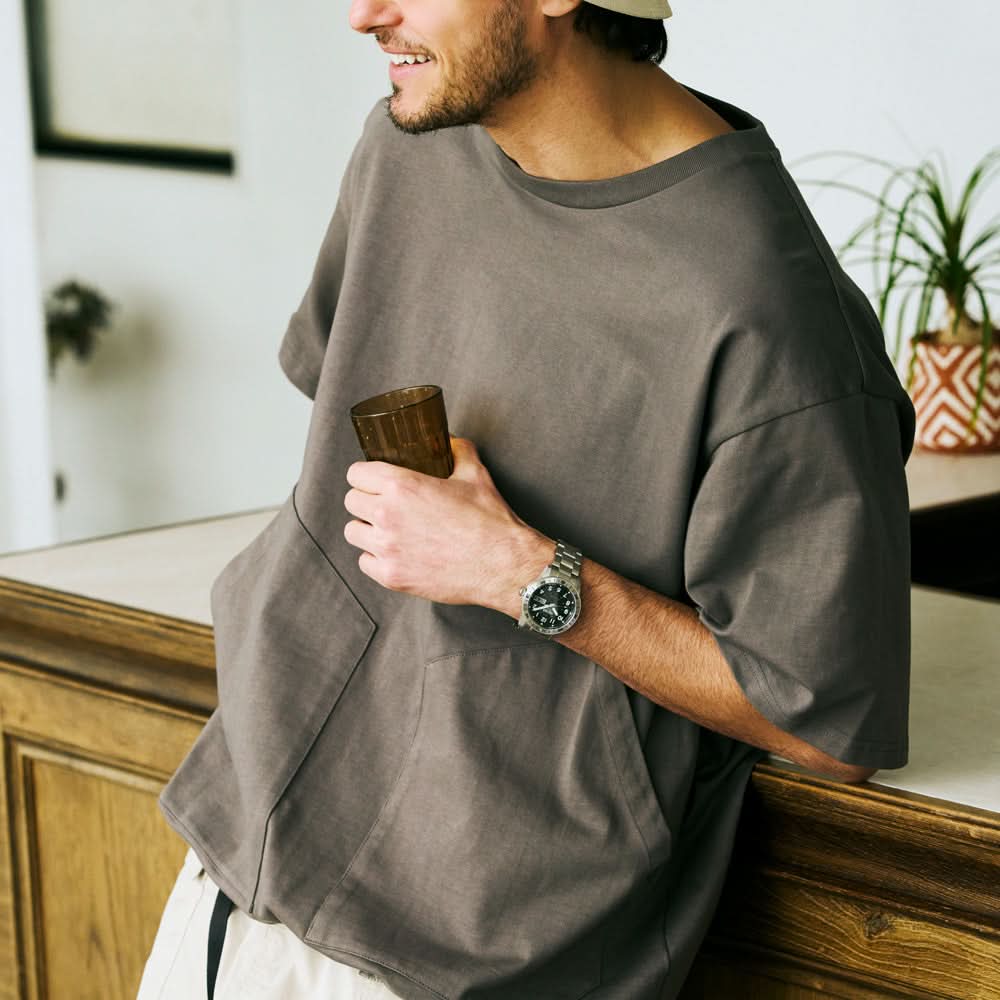 Man wearing a gray oversized shirt holding a wooden cup indoors with watch on his wrist.