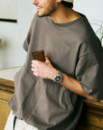 Man wearing a gray oversized shirt holding a wooden cup indoors with watch on his wrist.