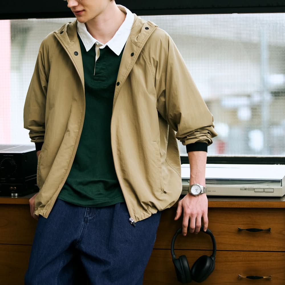 Person wearing a beige jacket over a green shirt with headphones on a wooden surface with white dial watch.