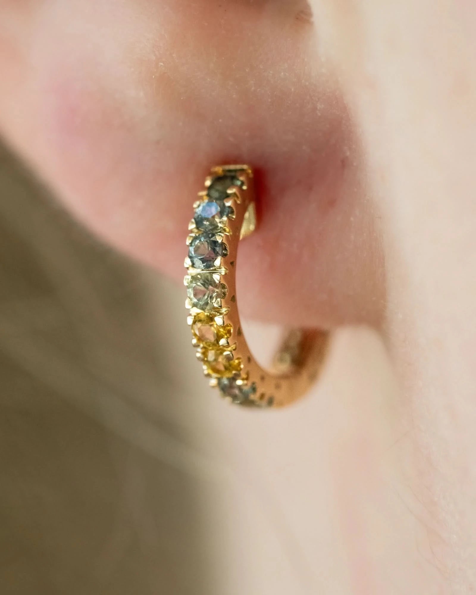 A close-up shot of a gold hoop earring adorned with small round gemstones in yellow, green, and blue tones, worn on a person's ear.