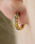 A close-up shot of a gold hoop earring adorned with small round gemstones in yellow, green, and blue tones, worn on a person's ear.