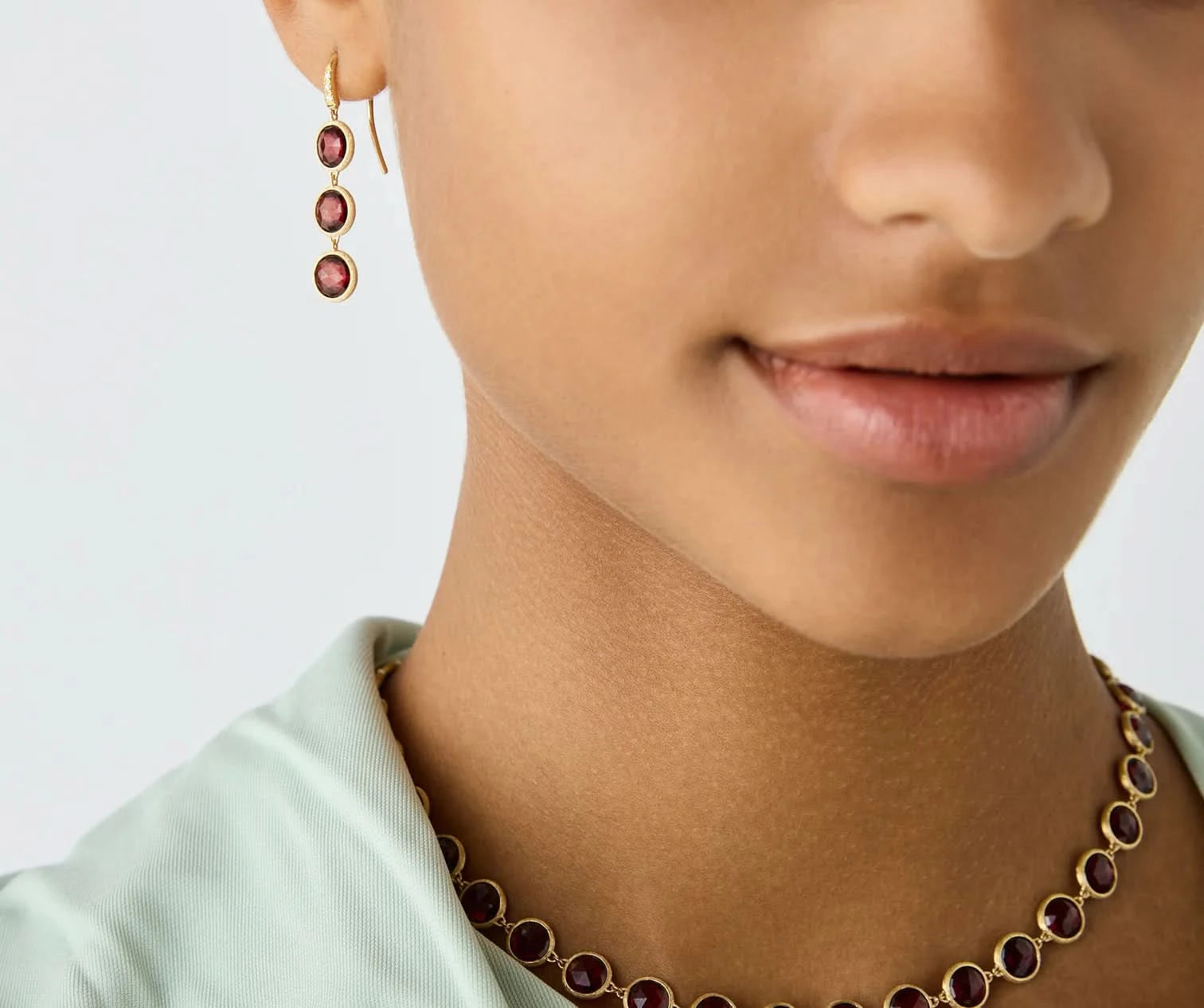 Close-up of a model wearing a gold necklace and matching drop earring, both featuring round deep red gemstones, against a clean light gray background.