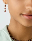 Close-up of a model wearing a gold necklace and matching drop earring, both featuring round deep red gemstones, against a clean light gray background.