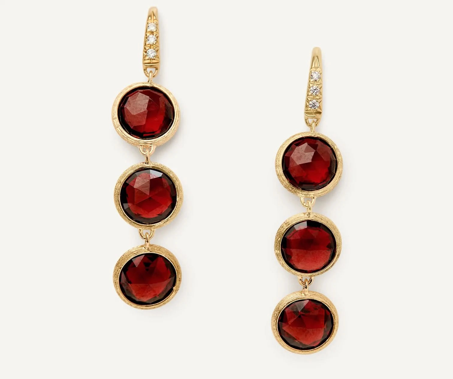 A pair of gold drop earrings featuring three faceted round red gemstones in each earring, accented with small clear stones along the top hooks, set against a plain off-white background.
