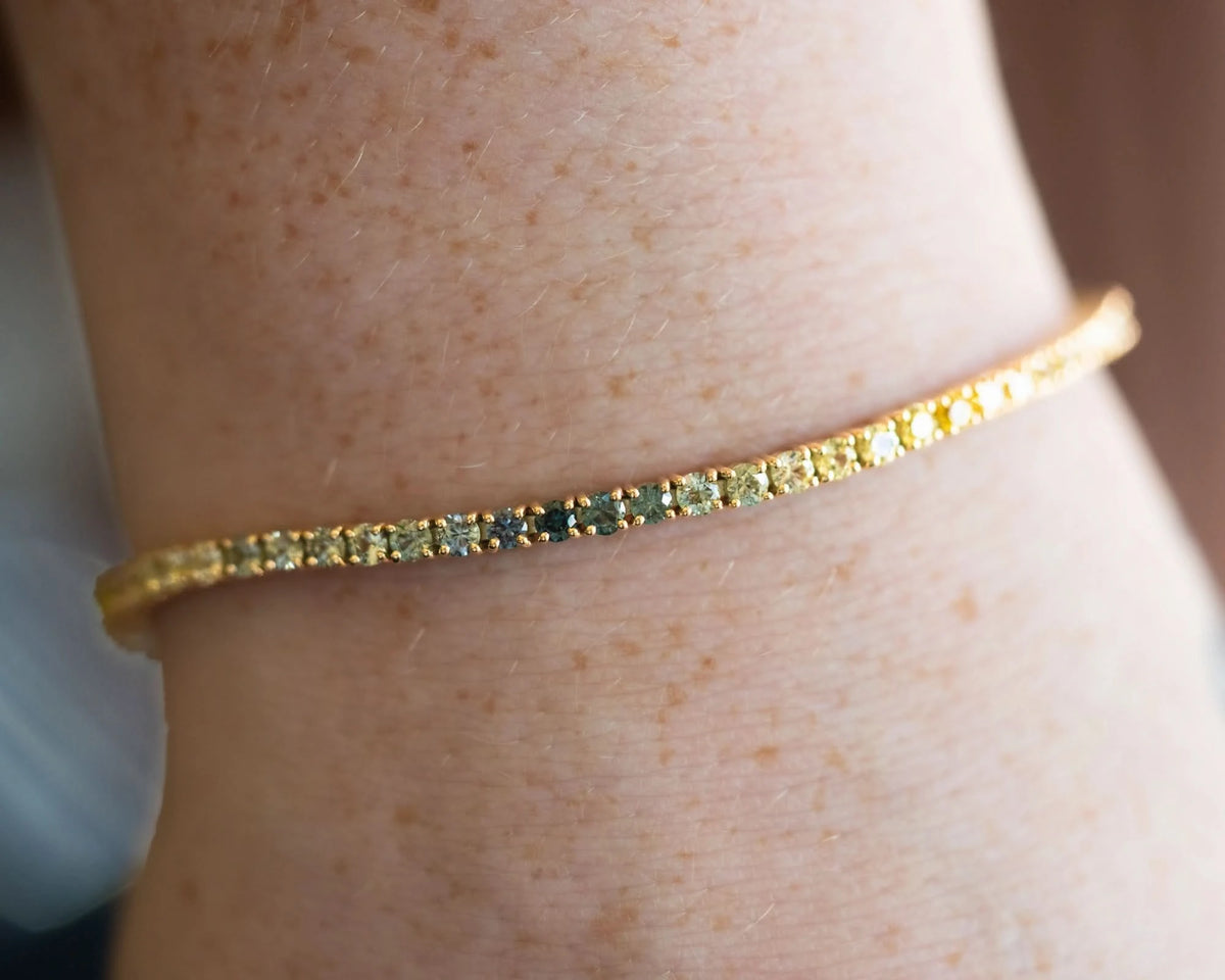 A close-up of a gold tennis bracelet with small yellow and green stones, worn on a person's wrist.