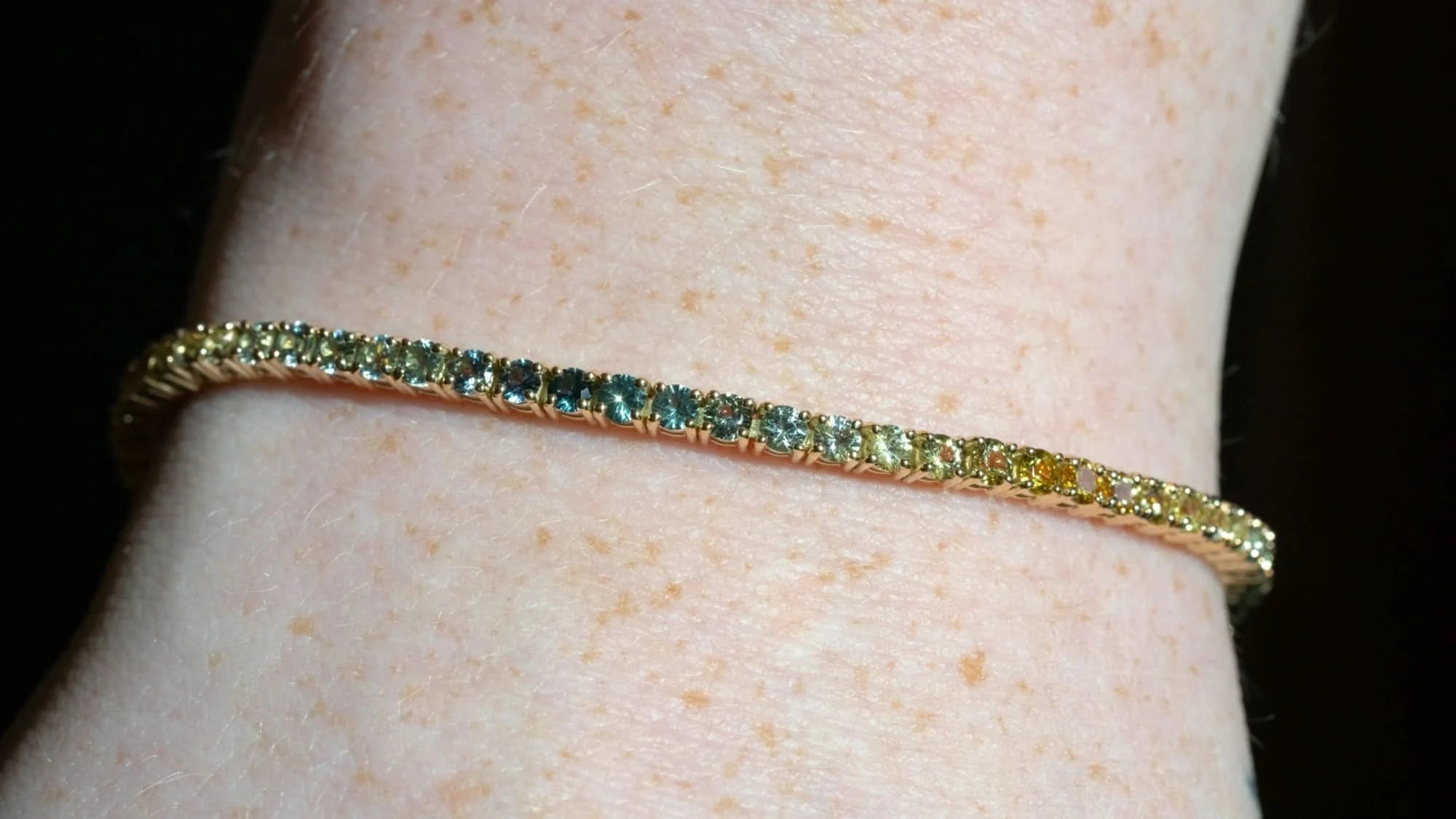 A close-up of a gold tennis bracelet with a row of small colored stones, worn on a person's wrist against a dark background.