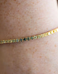 A close-up of a gold tennis bracelet with small yellow and green stones, worn on a person's wrist.
