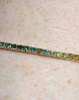 A close-up of a gold tennis bracelet with a row of small colored stones, worn on a person's wrist against a dark background.
