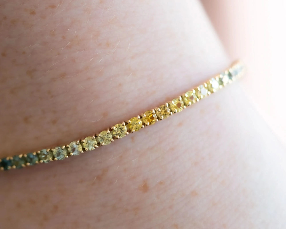 A close-up of a delicate gold tennis bracelet with alternating yellow and green stones, worn on a person's wrist.