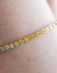 A close-up of a delicate gold tennis bracelet with alternating yellow and green stones, worn on a person's wrist.