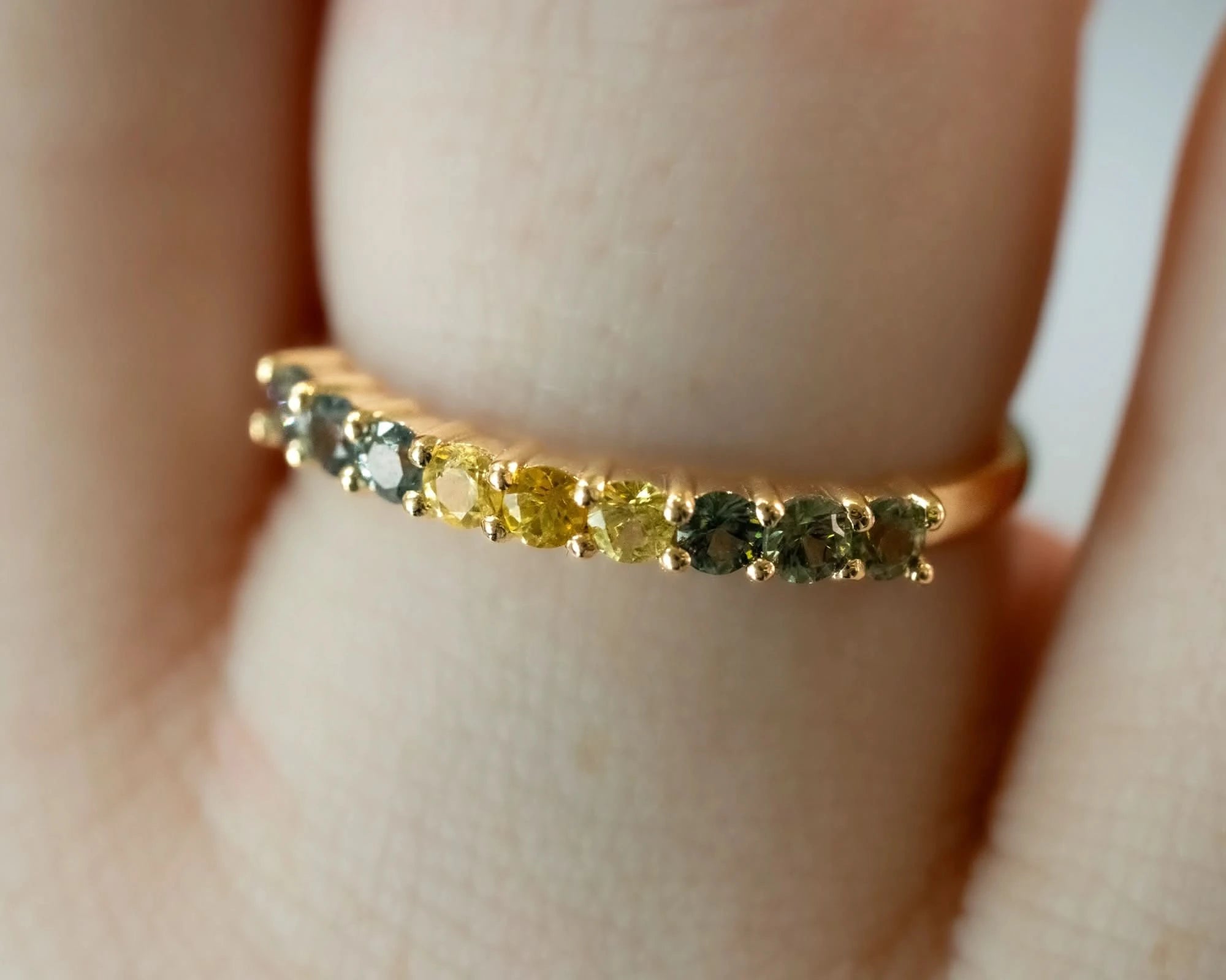 A close-up of a gold ring worn on a finger, featuring a row of small round gemstones in yellow, green, and gray tones.