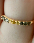 A close-up of a gold ring worn on a finger, featuring a row of small round gemstones in yellow, green, and gray tones.