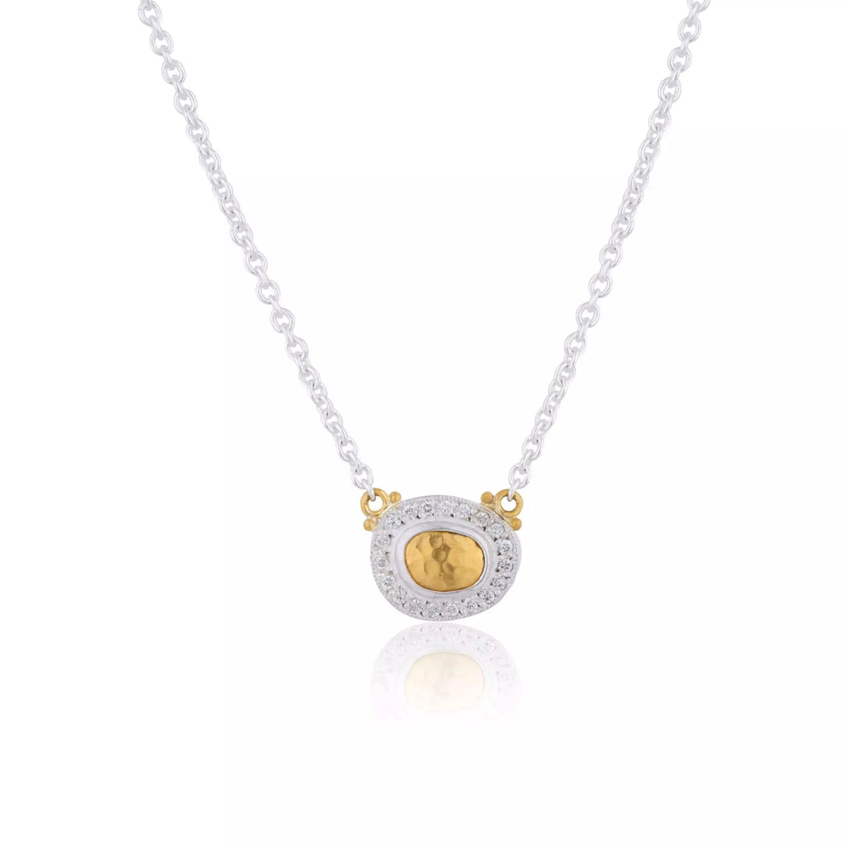 A delicate silver chain necklace featuring an oval pendant with a halo of small clear stones and a central gold-toned oval hammered metal, displayed on a clean white background with a soft reflection below.