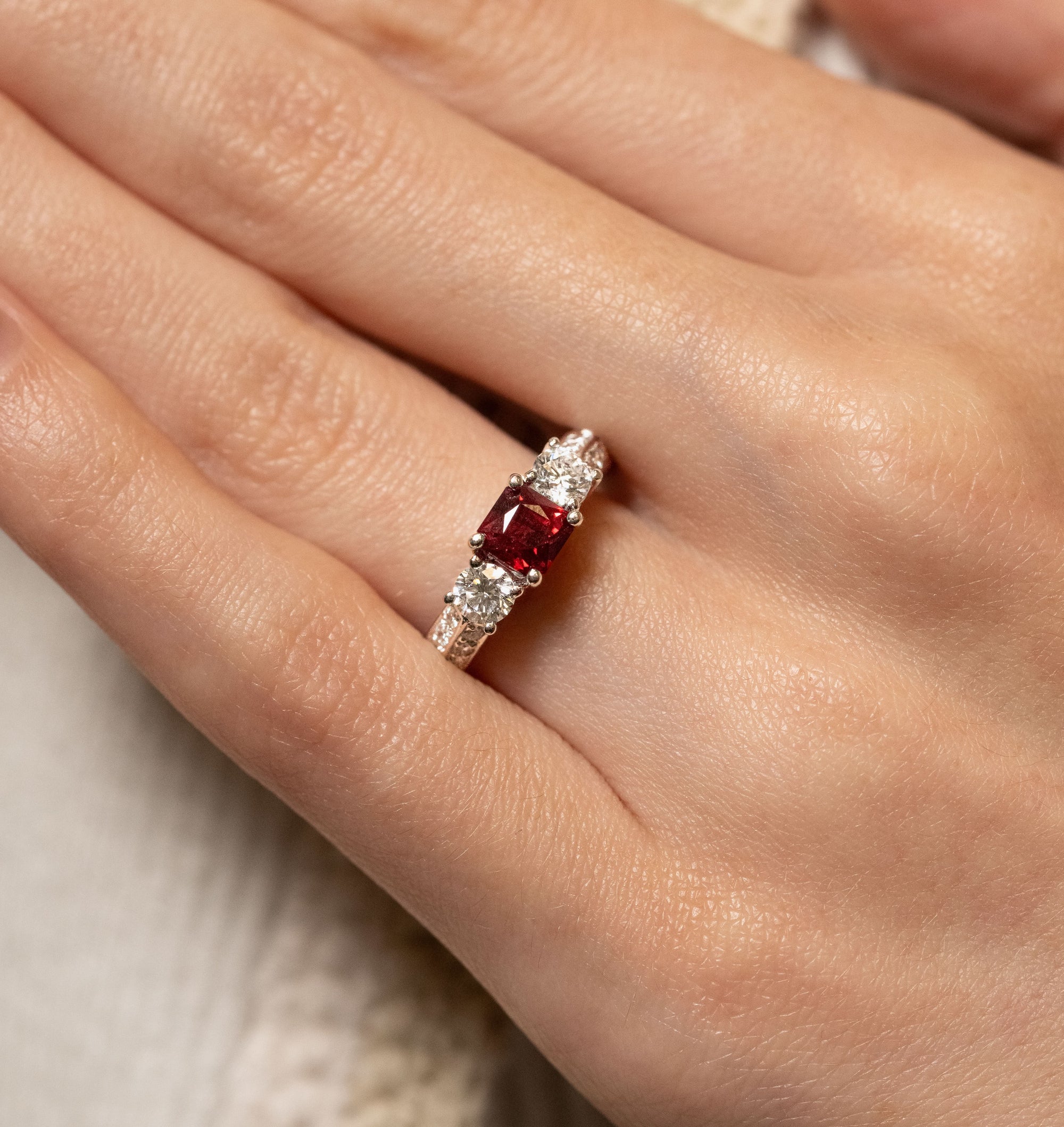 A close-up of a hand wearing a ring with a central square-cut red gemstone and clear side stones on a platinum band.