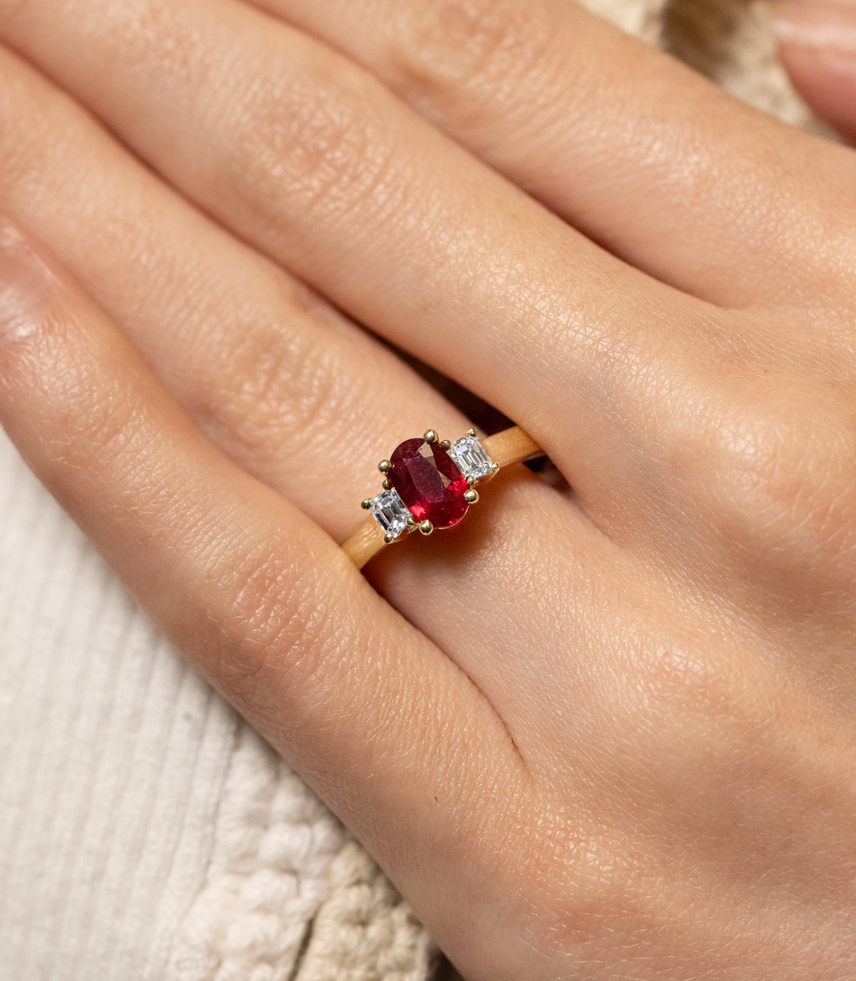 A close-up of a hand wearing a gold ring with an oval red center stone and two small clear side stones, resting on a textured cream fabric background.