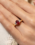 A close-up of a hand wearing a gold ring with an oval red center stone and two small clear side stones, resting on a textured cream fabric background.