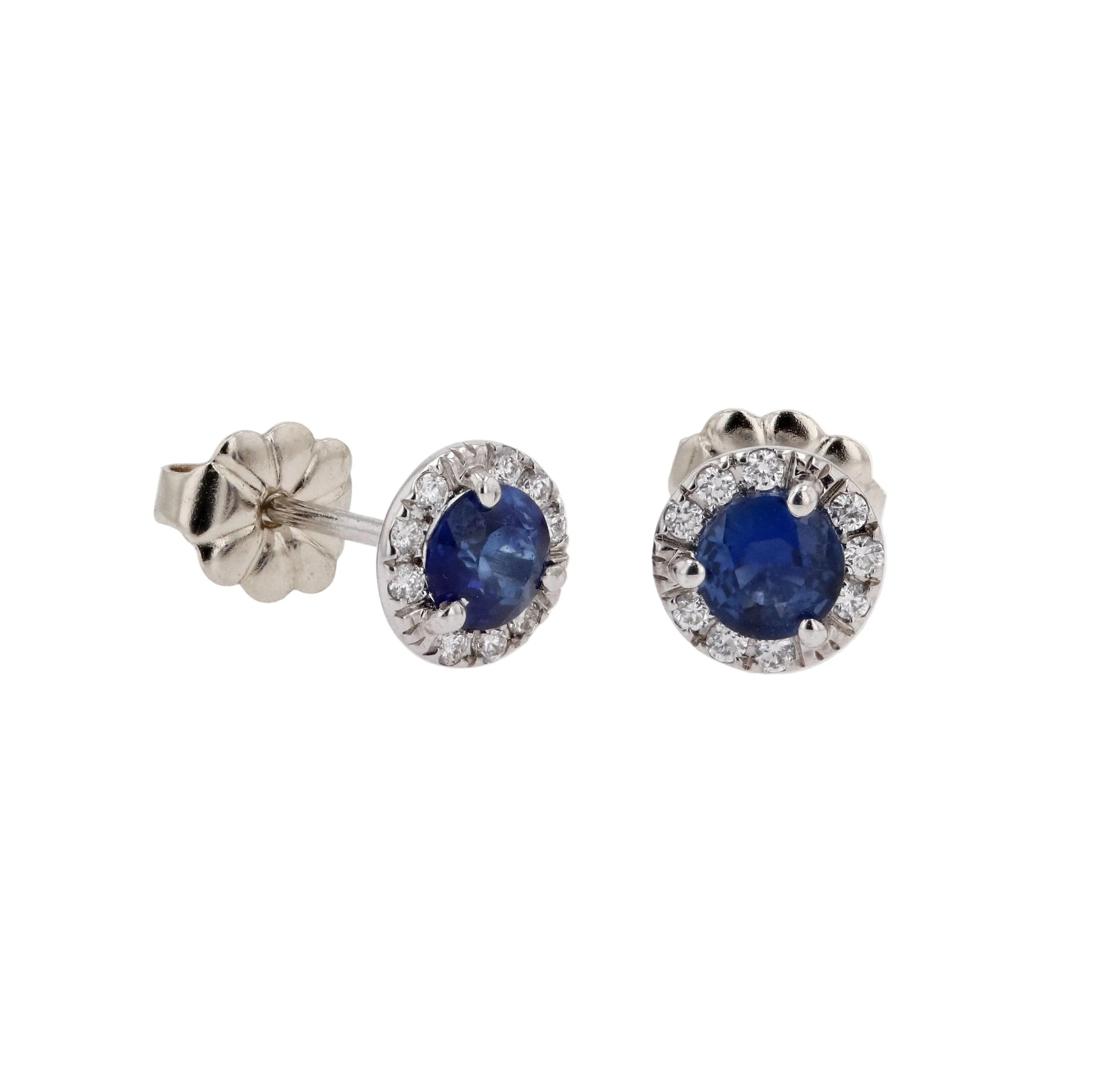 A pair of 14k white gold halo stud earrings featuring vibrant 4mm round blue sapphires encircled by a shimmering border of brilliant-cut white diamonds, resting on a  white backgound.
