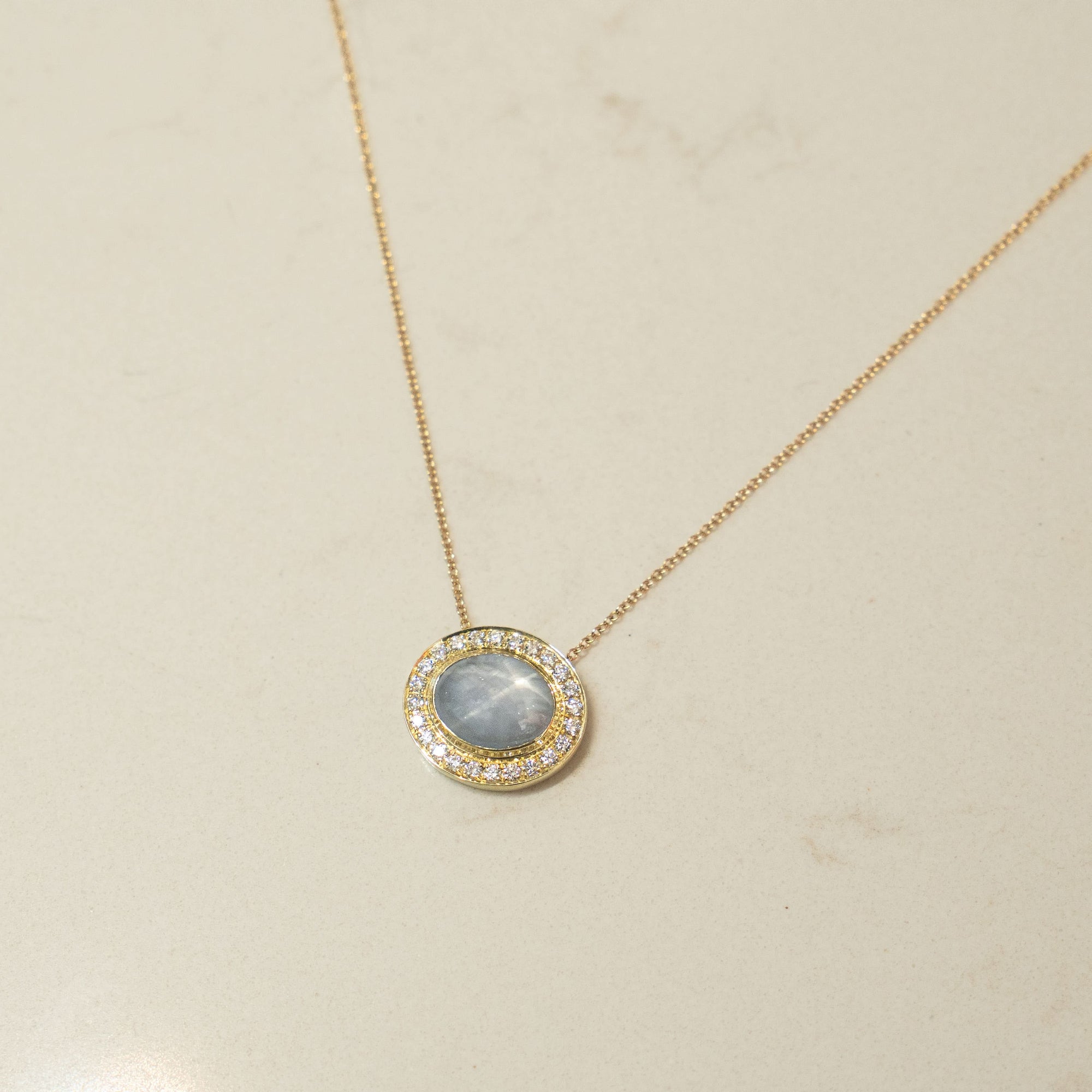 Gold necklace with an oval blue star sapphire  and diamond pendant on a beige background