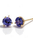 A pair of gold stud earrings with round-cut deep purplish blue tanzanite gemstones, shown on a clean white background with soft reflections.