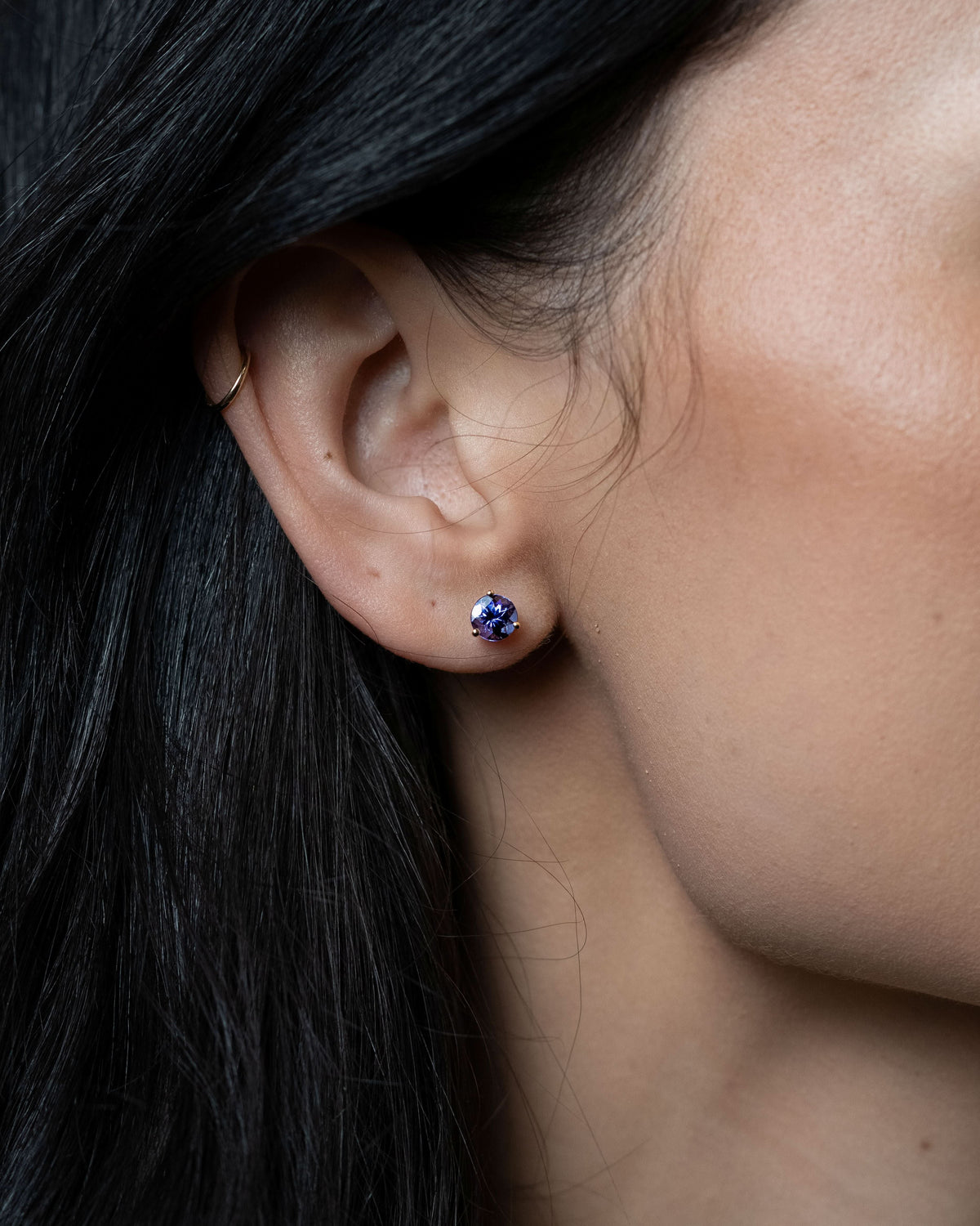 A close-up of a person's ear wearing a small round stud earring with a blue center set in a gold-tone bezel, with dark hair framing the side of the face.