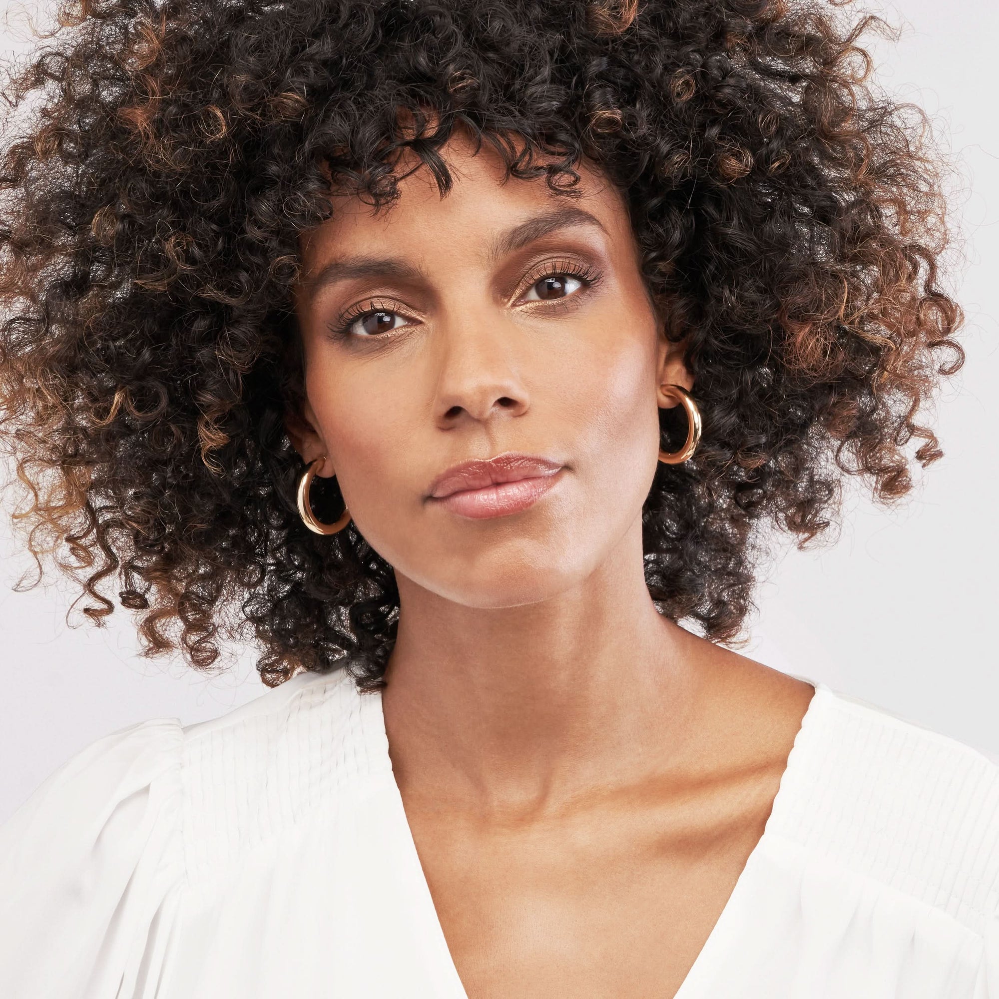 Woman with curly hair wearing gold hoop earrings against a white background