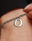 A close-up of a silver chain anklet with a polished oval letter 'O' charm worn on a person's ankle, with fingers holding the chain and a black ribbed sock visible at the top.