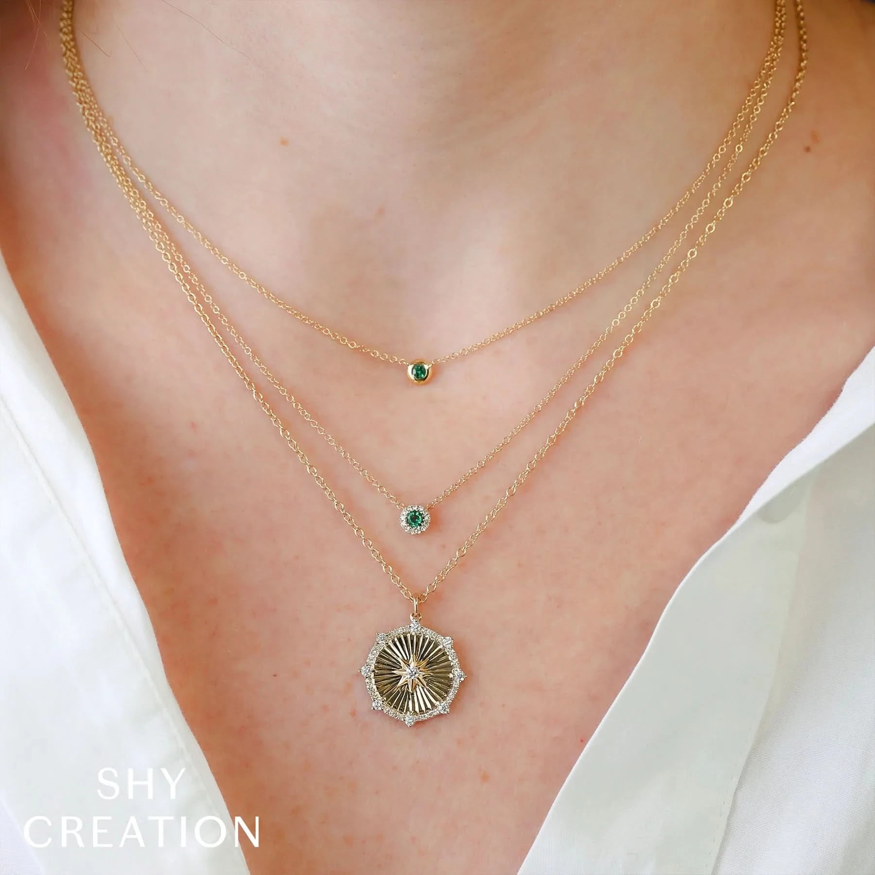 Two gold necklaces with green gemstones and one with a medallion on a person wearing a white top, branded 'SHY CREATION'.