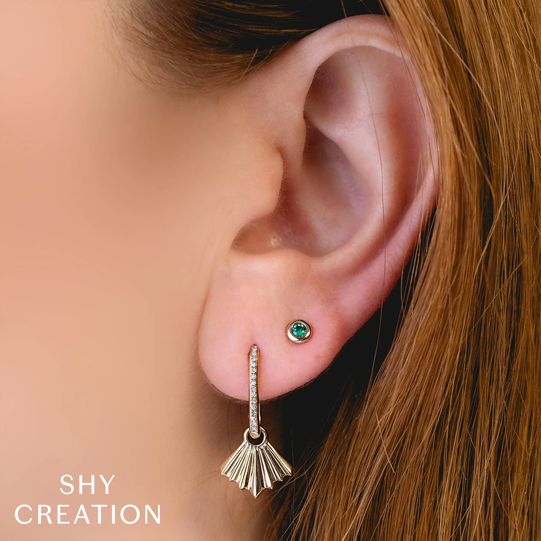 Close-up of an ear wearing a gold earring with a green gemstone, branded 'SHY CREATION'.