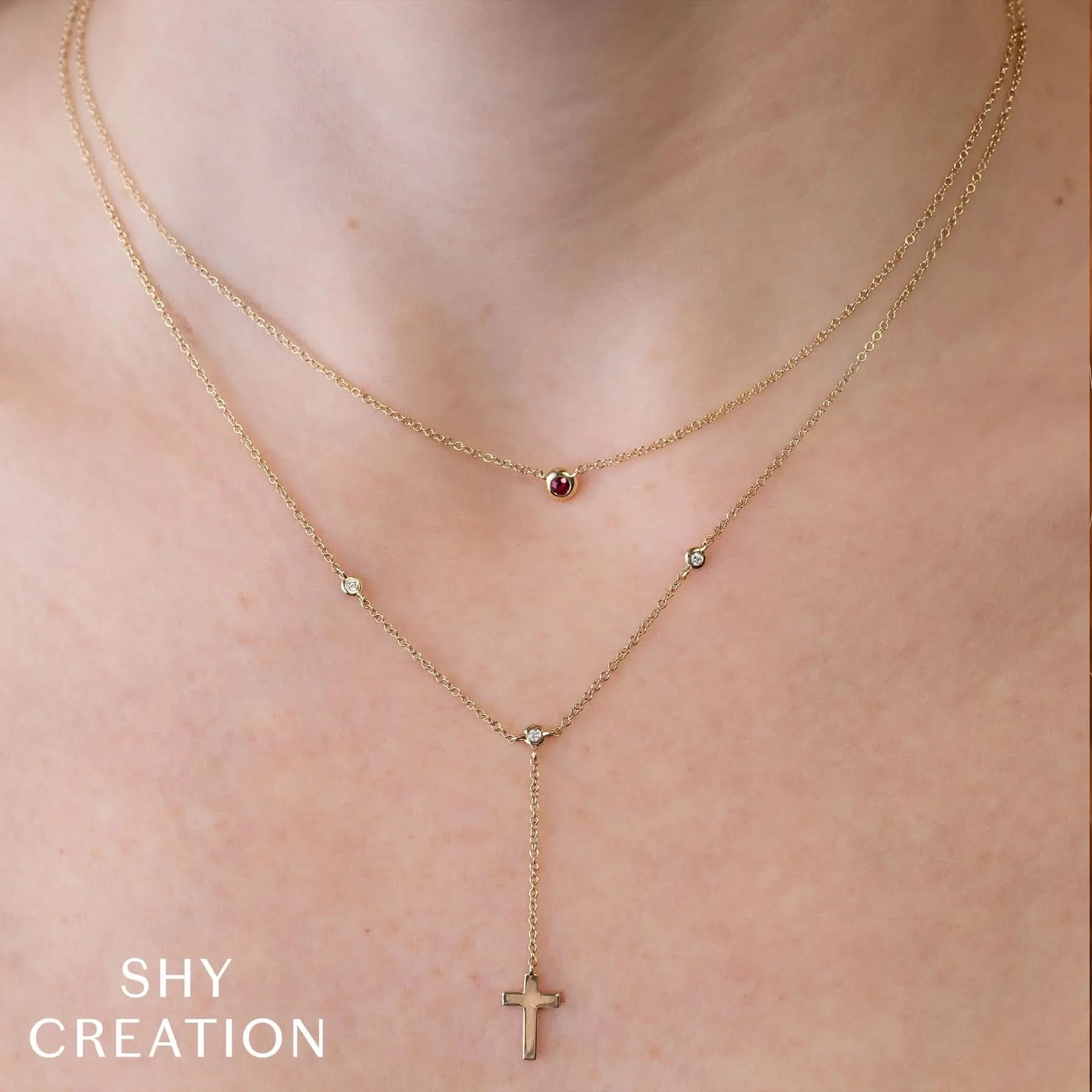 Gold necklace stack one with  red bezel and one with a cross pendant on a person's neck, branded 'SHY CREATION'.