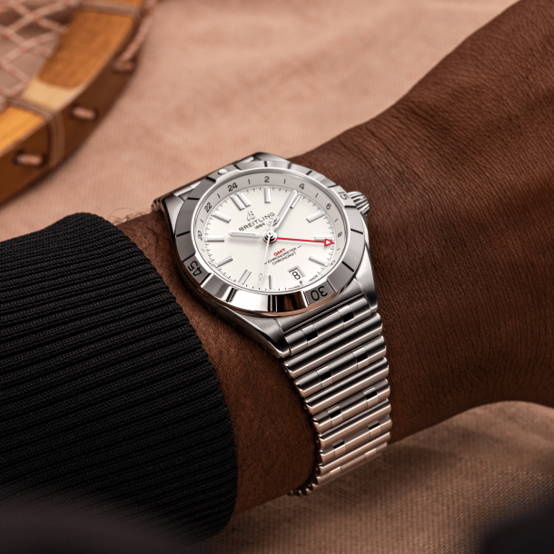 A wrist wearing a silver-toned Breitling watch with a white dial and date window, photographed against a warm brown  background.