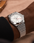 A wrist wearing a silver-toned Breitling watch with a white dial and date window, photographed against a warm brown  background.