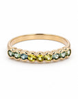 A gold ring featuring a row of faceted gemstones in shades of green, blue, and yellow, set against a clean white background.