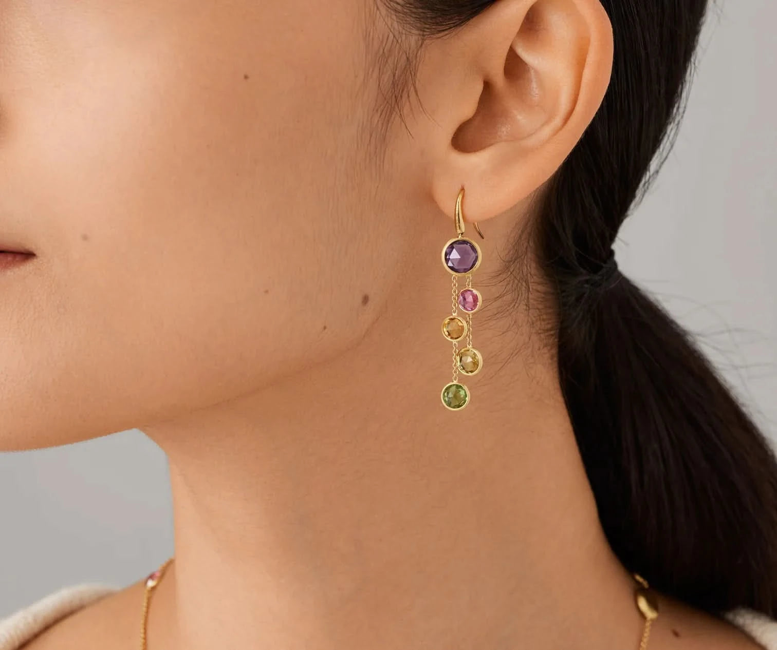 Close-up of a person wearing a gold earring with colored gemstones on a neutral background