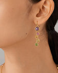 Close-up of a person wearing a gold earring with colored gemstones on a neutral background