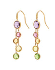 A pair of gold dangle earrings featuring a vertical cascade of round-cut gemstones in purple, pink, yellow, and green, set in gold hooks against a light cream background.