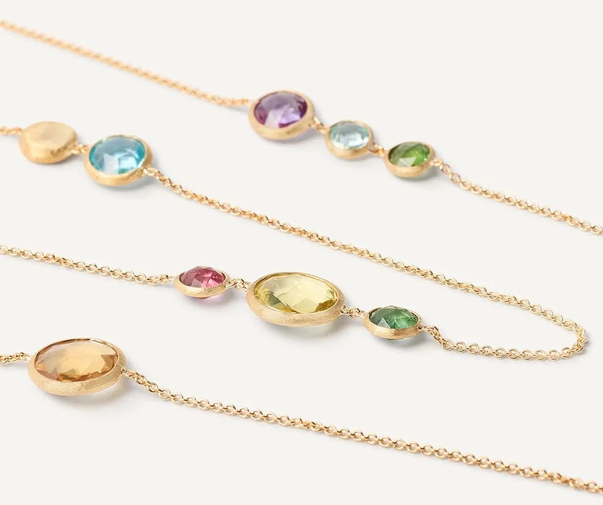 Three delicate gold chain necklaces with oval and round multicolored gemstones, arranged diagonally on a clean white background.