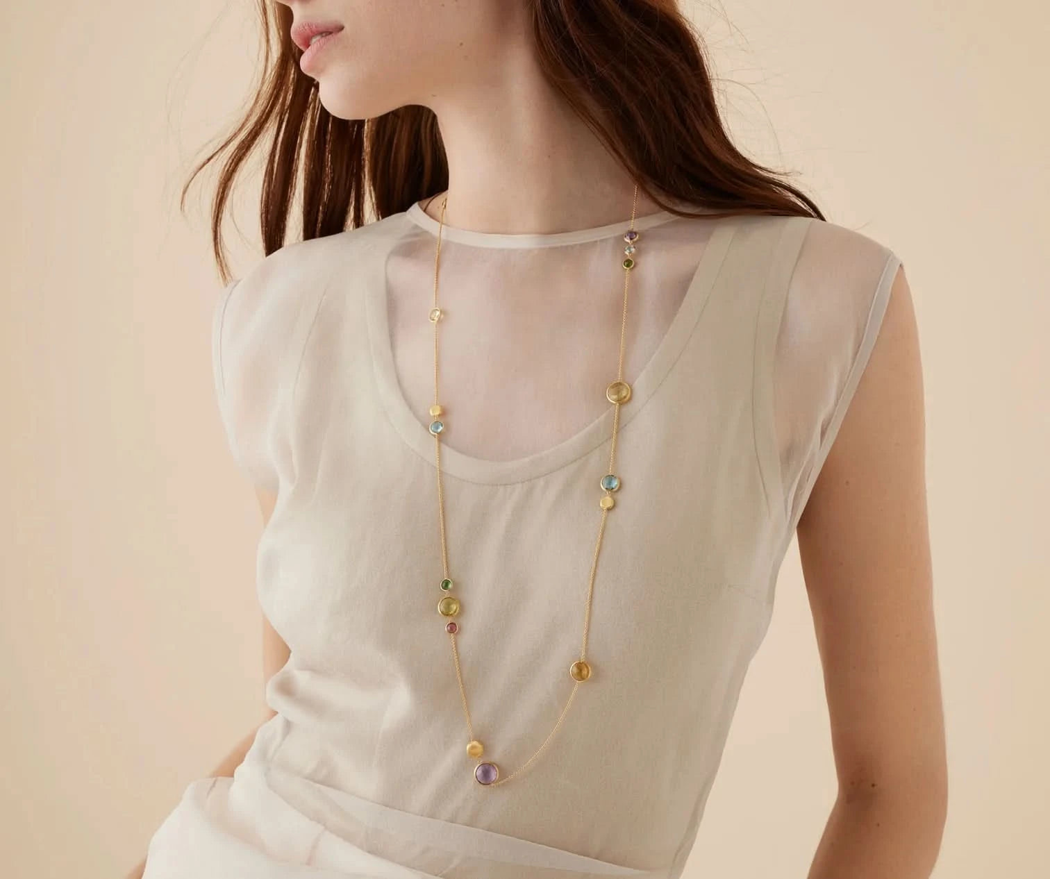 A woman wearing a long gold-toned beaded necklace with small multicolored beads over a sheer cream top, photographed against a soft beige background.