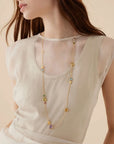 A woman wearing a long gold-toned beaded necklace with small multicolored beads over a sheer cream top, photographed against a soft beige background.