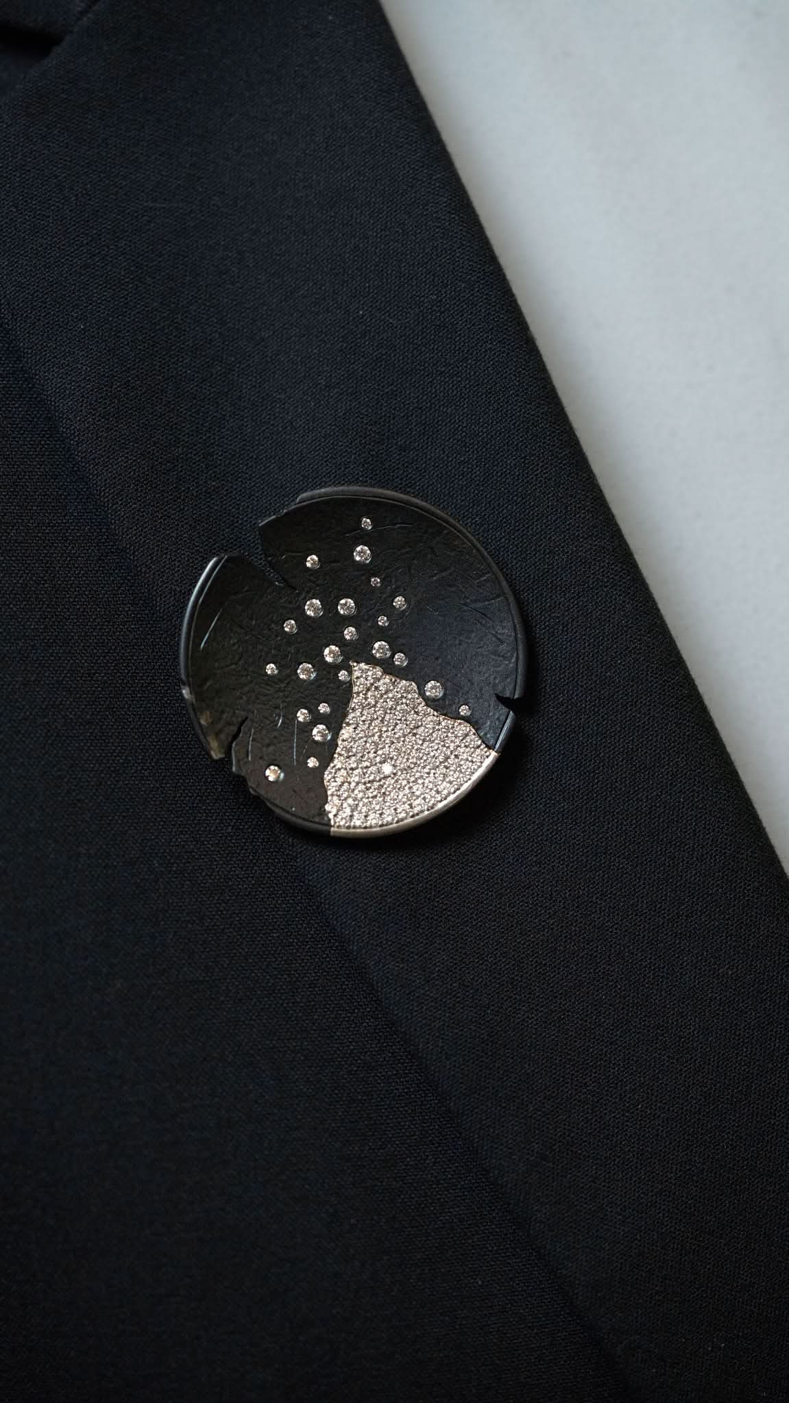 Logan James black steel and 18K white gold diamond brooch worn on black blazer lapel close-up
