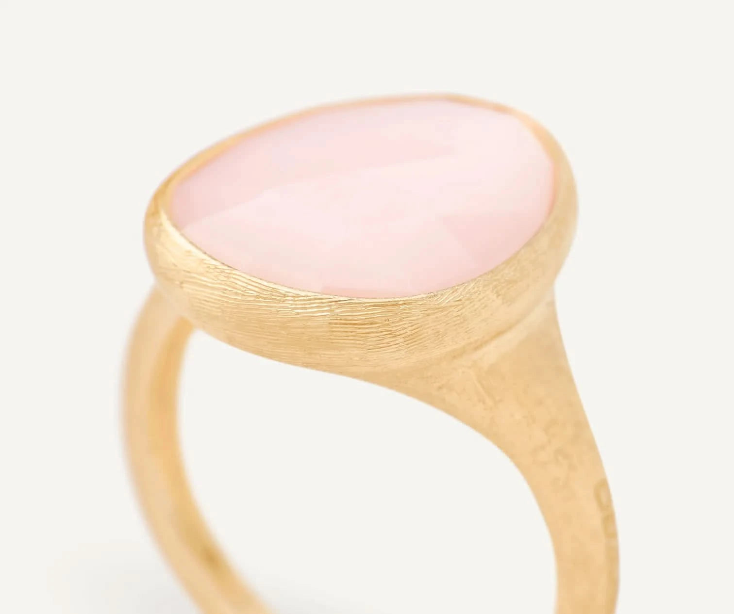 A close-up shot of a gold signet ring featuring a smooth, oval-shaped pale pink stone, set against a clean, light off-white background.