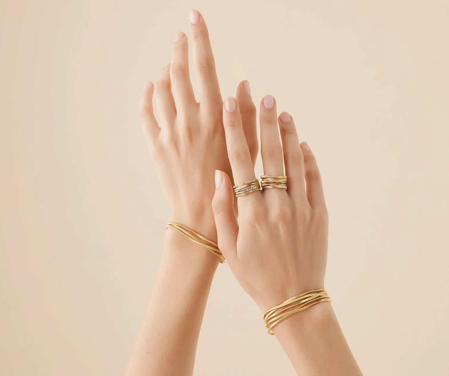 A pair of hands wearing multiple delicate gold rings and matching gold bangle bracelets, set against a solid warm beige background.