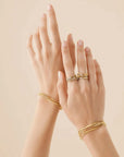 A pair of hands wearing multiple delicate gold rings and matching gold bangle bracelets, set against a solid warm beige background.
