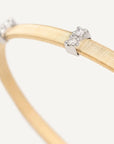 A close-up of a gold bangle bracelet with silver-toned end caps, each set with small clear gemstones, on a light off-white background.