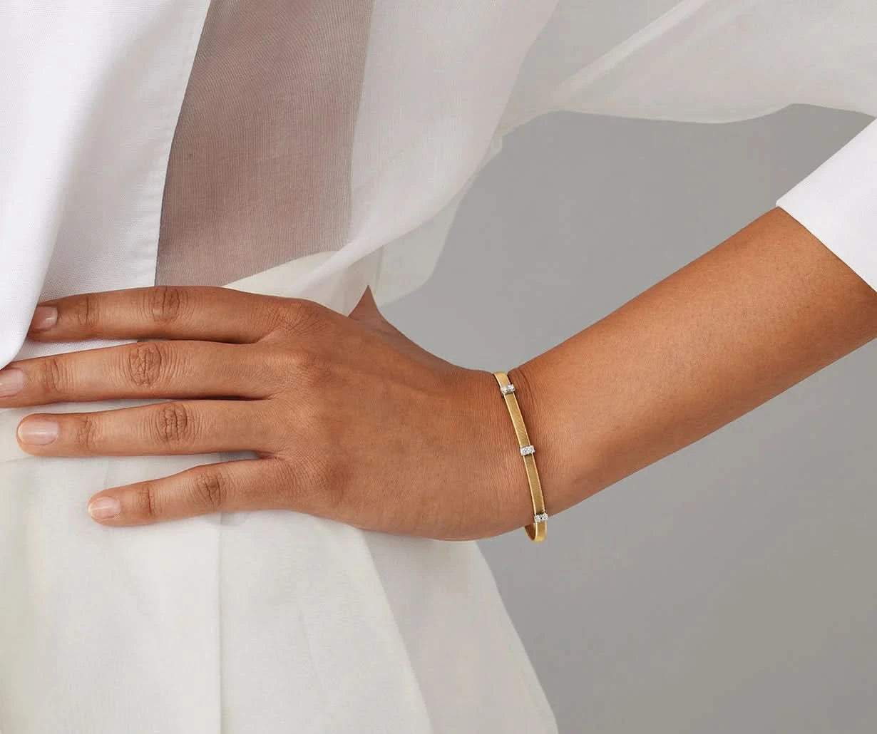 A close-up of a person's hand and wrist wearing a slim gold bangle bracelet with small silver-toned rectangular accents, styled with a white draped garment against a soft gray background.
