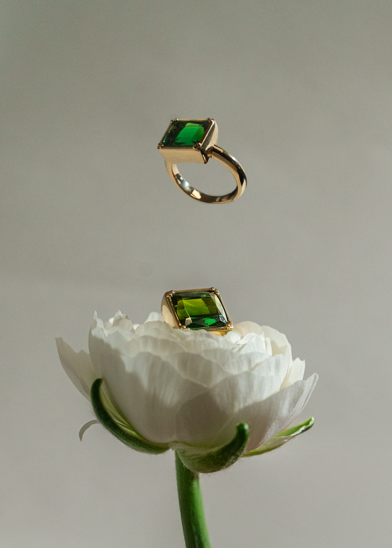 Two gold rings with rectangular green gemstones are shown against a soft gray background, one floating above and one resting on a white flower.