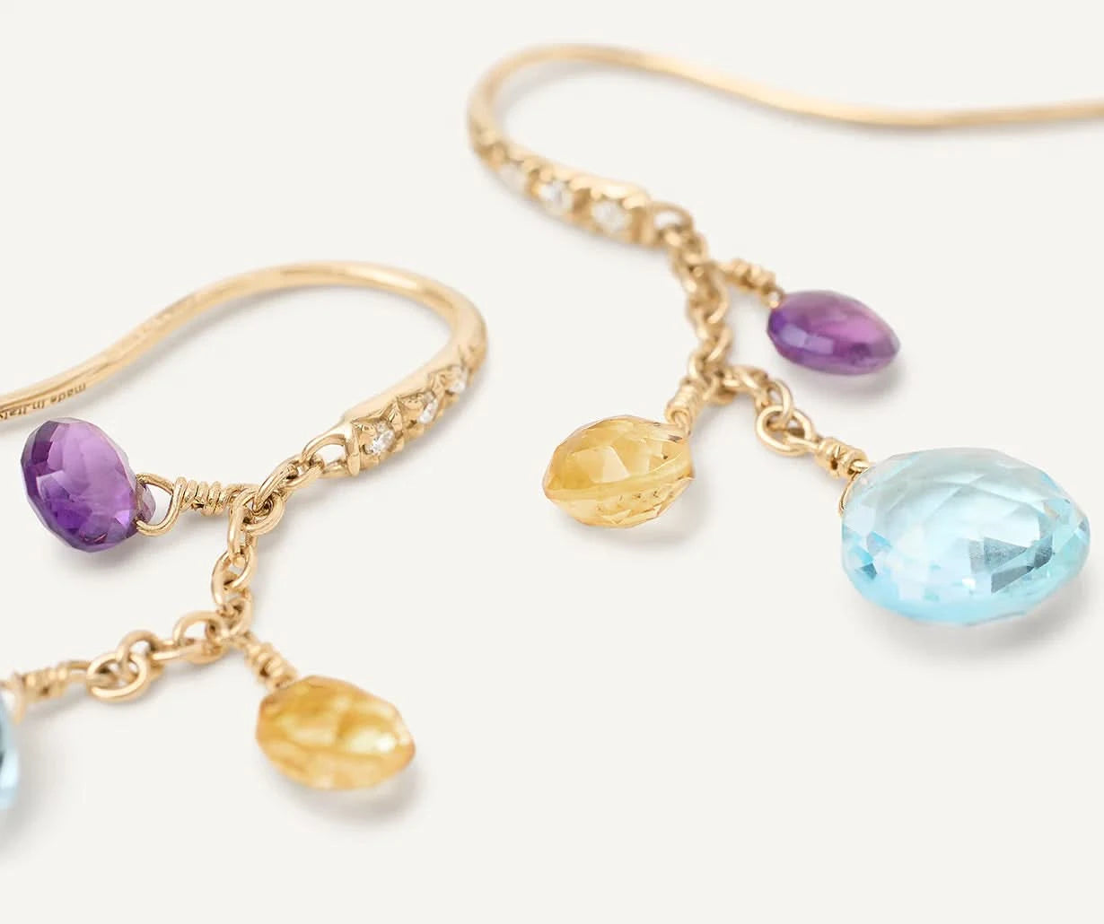 Gold dangle earrings with faceted gemstone charms in light blue, purple, and yellow, shown on a clean off-white background.