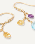 Gold dangle earrings with faceted gemstone charms in light blue, purple, and yellow, shown on a clean off-white background.