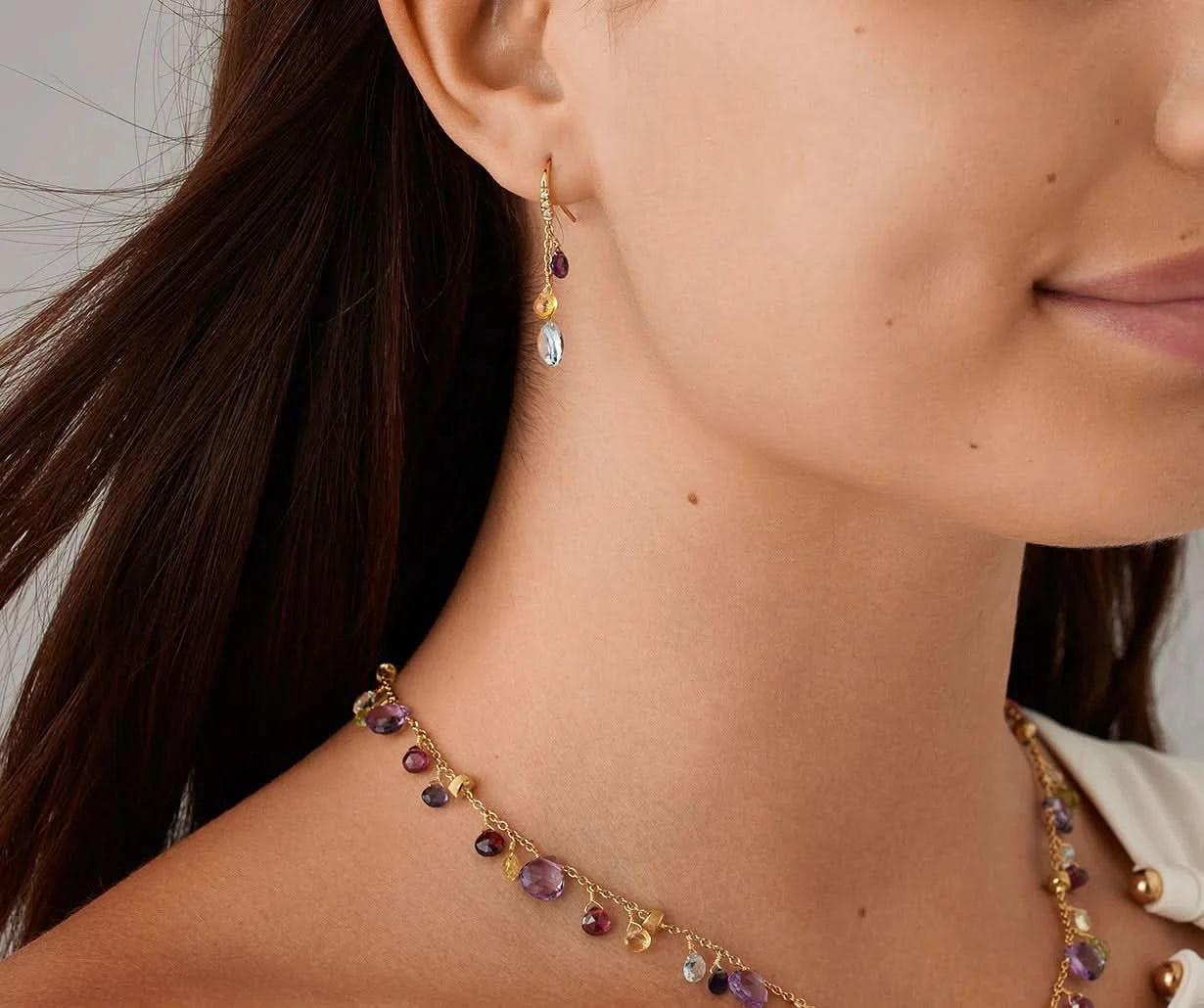 Close-up of a woman wearing a gold gemstone necklace and matching drop earring with small purple and pale blue gemstones